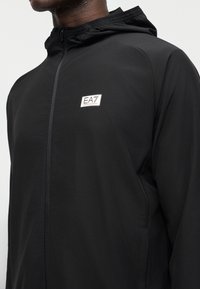 Μαύρο μπουφάν με φερμουάρ και κουκούλα που φοράει πρόσωπο, με μικρό λευκό λογότυπο EA7 Emporio Armani στο στήθος.