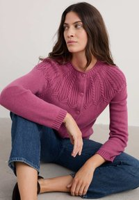 Cardigan de lana rosa con detalles texturizados en el yugo, presenta un cuello redondo y tres botones. Llevado con jeans de mezclilla azules.