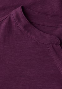 Camicetta viola con scollatura rotonda, tessuto morbido e motivo mélange sottile. Cuciture a vista lungo il colletto e le spalle.