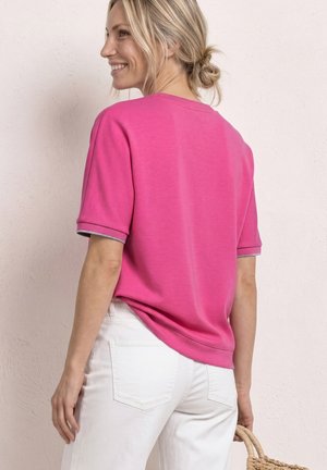 Femme souriante, portant un t-shirt rose à manches courtes et un pantalon blanc, tenant un sac tressé contre un mur clair texturé.