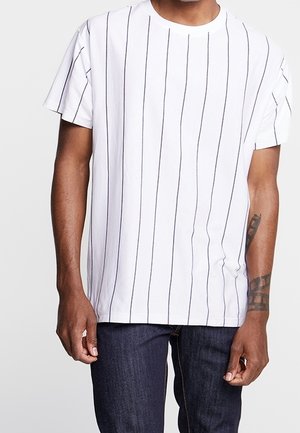 Mężczyzna ubrany w białą koszulkę z krótkim rękawem z cienkimi pionowymi czarnymi paskami oraz ciemnoniebieskie dżinsy, stojący na jednolitym tle.