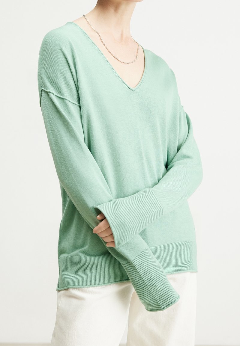 Personne portant un pull ample vert menthe à col en V avec de grandes poignets côtelées et un pantalon clair, debout les bras croisés.