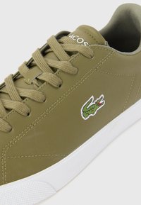 Sneakers verde-oliva com acabamento em pele lisa, apresentando solas e atacadores em borracha branca, e um logótipo de crocodilo bordado em verde.