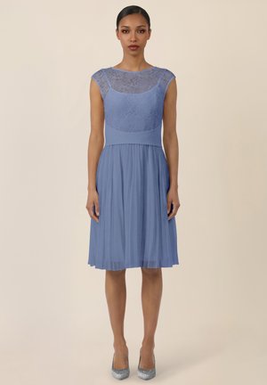 Woman wearing a knee-length, sleeveless blue dress with lace bodice and pleated skirt, standing against a plain beige background.