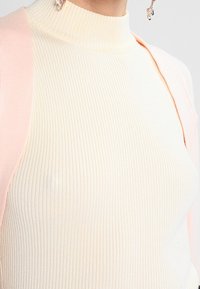 Suéter de cuello alto acanalado en beige cremoso, hecho de material suave. Presenta un cárdigan rosa pastel dispuesto sobre un hombro, añadiendo contraste.