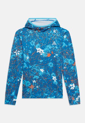 Camisa azul de manga larga con capucha, con patrones florales abstractos en naranja y blanco, y un pequeño logo triangular blanco en el pecho.