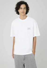 T-shirt en coton blanc à manches courtes, avec une petite impression noire, coupe décontractée et col rond. Porté avec un pantalon gris clair.
