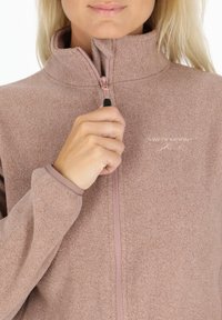 Polarowa kurtka w delikatnym mauve z wysokim kołnierzem, zamek z przodu i elastycznymi mankietami. Posiada haftowane logo na przedzie.