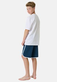 White cotton t-shirt and navy shorts with white trim, featuring a relaxed fit. The subject is barefoot, looking back over their shoulder.