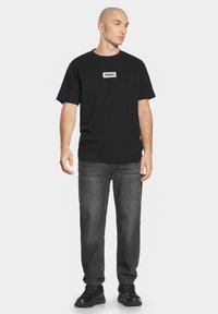 Czarny bawełniany t-shirt z białym prostokątnym logo "PROSTO.", w połączeniu z ciemnoszarymi dżinsami i czarnymi sneakerami, stojący na jednolitym tle.