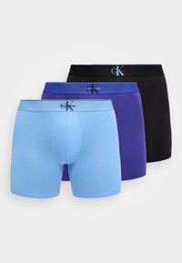 Τρία ζευγάρια αντρικά εσώρουχα boxer Calvin Klein σε ανοιχτό μπλε, μωβ και μαύρο, τοποθετημένα το ένα πίσω από το άλλο σε λευκό φόντο.