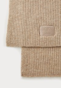 Écharpe beige côtelée en laine douce, présentant une finition texturée et un petit logo de marque embossé sur une étiquette rectangulaire.