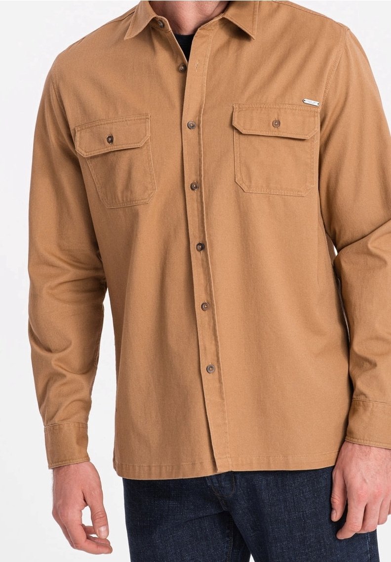 Man draagt een beige overhemd met twee borstflapzakken en donkerblauwe jeans, te zien van nek tot heupen.