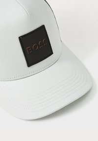 Hvitt baseball-cap med en strukturert fasong, prydet med en svart rektangulær lapp med "BOSS" stemplet i kobber. Teksturert stoff.