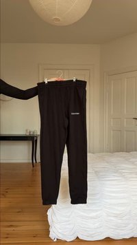 Melni Calvin Klein sporta bikses, kas pakārtas uz pakaramā minimālistiskā guļamistabā ar koka grīdu, baltu gultu un slēgtām durvīm fona.