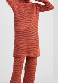 Personne portant un pull en tricot rayé orange et noir avec un pantalon assorti, debout les bras levés devant un fond uni.
