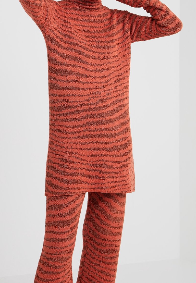 Personne portant un pull en tricot rayé orange et noir avec un pantalon assorti, debout les bras levés devant un fond uni.