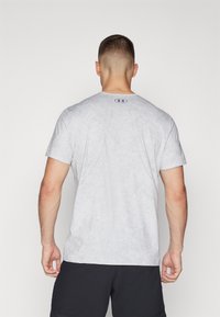 Camiseta deportiva de color gris claro con textura suave, mangas cortas y detalle perforado en la parte posterior. Presenta un logo circular en el cuello.