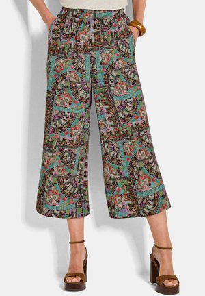 Frau trägt bunte, weit geschnittene, verkürzte Hosen mit Blumen- und geometrischen Mustern, braune Plateau-Sandalen und ein Armband am Handgelenk.