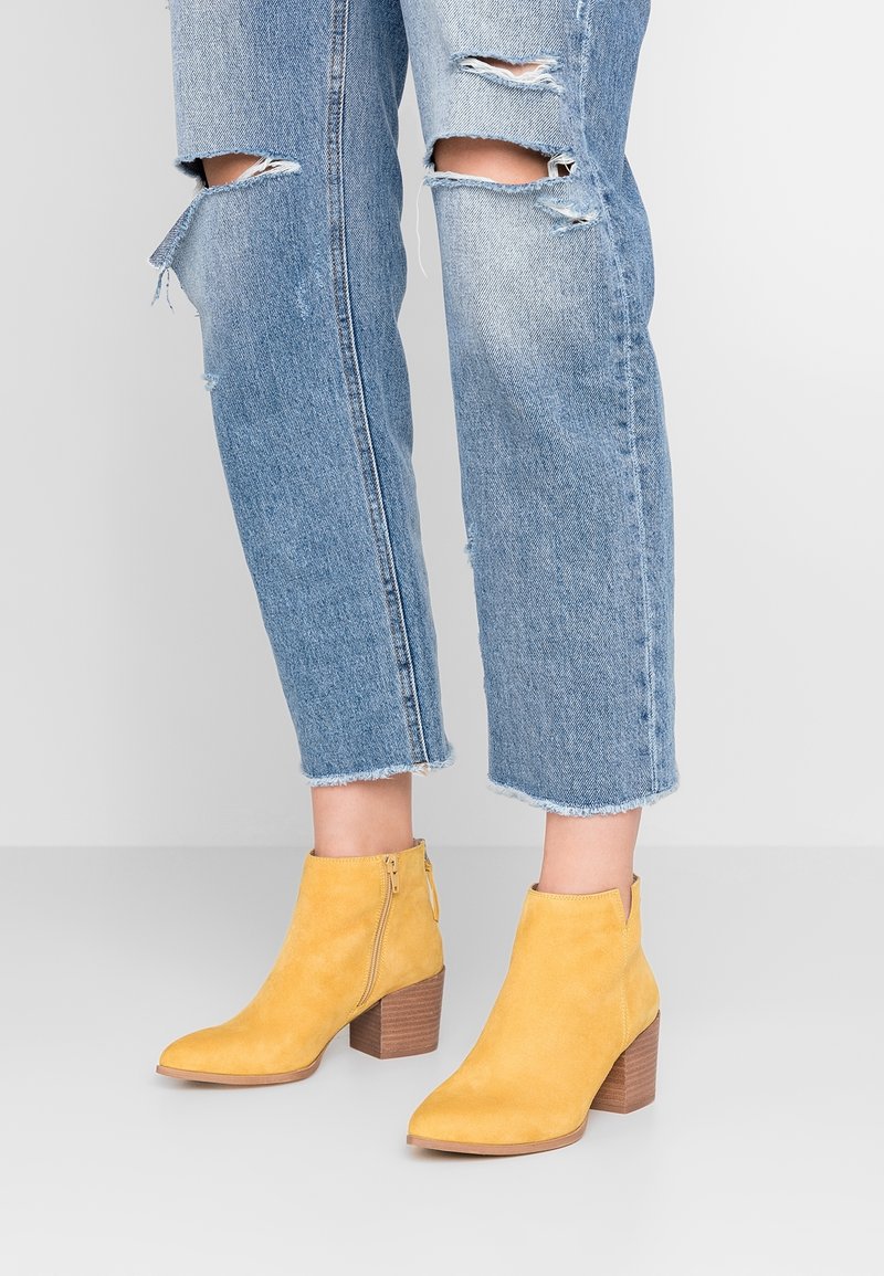 Botas de tobillo de ante amarillo con punta afilada y tacón cuadrado. Cuentan con una cremallera lateral y un tacón de madera texturizada. Combinadas con jeans azul desgastados.