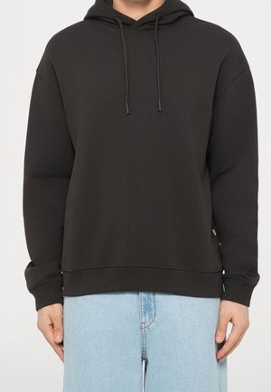 Personne portant un sweat à capuche noir uni avec des cordons et un jean bleu clair, montrée des épaules aux hanches sur un fond blanc.