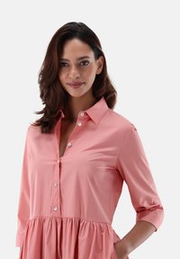 Vestito rosa con bottoni, dotato di colletto, con un corpo aderente e una gonna ampia e plissettata. Maniche lunghe arrotolate fino ai gomiti. Tessuto liscio.