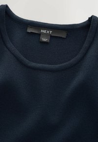 Sötétkék kötött pulóver, kerek nyakkal. A címkék láthatóak, és a "NEXT" márkát, valamint a "KÍNAI TERMÉK" származást jelzik, sima textúrával.