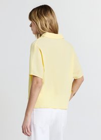 Chemise jaune à manches courtes avec finition texturée et col pointu, associée à un pantalon blanc. Accent sur le tissu léger et la coupe décontractée.