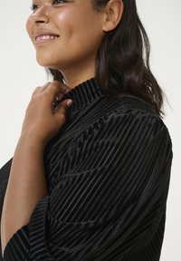 Haut en tissu noir à côtes avec un col haut. Présente des rayures verticales et une surface texturée. Des manches courtes et bouffantes accentuent le design.