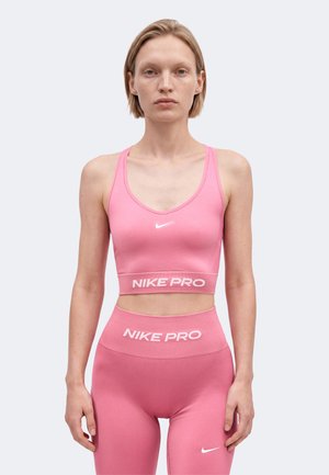 Femme portant un soutien-gorge de sport rose Nike Pro assorti et un legging taille haute, debout devant un fond clair uni.
