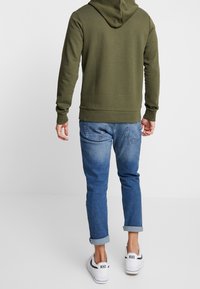 Olivgrön hoodie med snörning i huva, ribbade ärmslut och nederkant, parat med uppvikta blå jeans och vita sneakers.