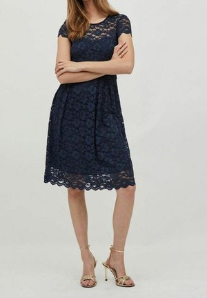 Kvinde iført en marineblå blonderkjole med korte ærmer, armene krydsede, og gyldne sandaler med høje hæle og stropper mod en neutral baggrund.