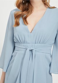 Žena v světle modrých šatech s výstřihem do V, 3/4 rukávy a vázaným textilním páskem v pase.