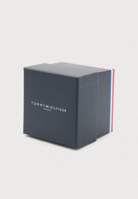 Tumši zilā krāsā Tommy Hilfiger pulksteņu kastīte ar baltu logo un sarkano, balto un zilo svītru malām uz gaiša fona.