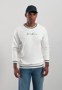 Vit sweatshirt med gröna detaljer på kragen och ärmsluten, med en kursiv "Ombree"-design. Matchas med blå jeans.