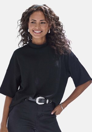 Lächelnde Frau mit lockigem Haar trägt schwarzes, strukturiertes Kurzarmoberteil, hoch taillierte schwarze Hose, silberne Creolen und Armband, Hand in die Hüfte gestützt.