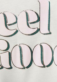 Tessuto bianco con testo audace e multicolore che recita "feel good". Le lettere sono spesse con contorni metallici texturizzati in turchese e bronzo.