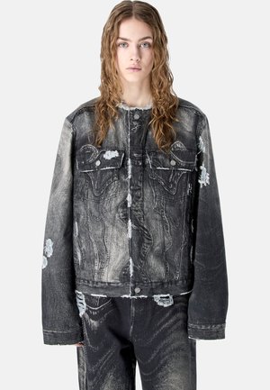 Young person with long curly hair wearing a distressed black denim jacket and matching pants, standing against a light background.