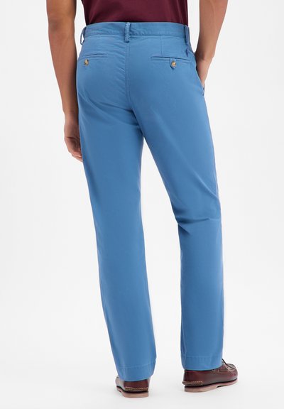 Jasnoniebieskie spodnie chinos o prostym kroju nogawki, z zapięciem na guzik w tylnej kieszeni i gładkim bawełnianym wykończeniem. Noszone z brązowymi butami.