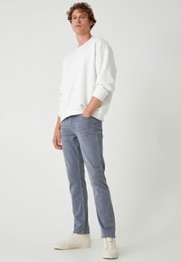 Jeans ajustados de color gris claro combinados con un sudadera blanca de cuello redondo y zapatillas beige, contra un fondo liso.