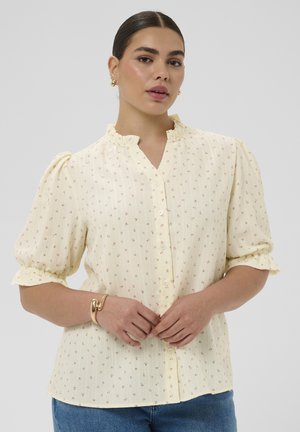 Žena v krémovej blúzke s malým kvetinovým vzorom, nafúknutými rukávmi, volánovým golierom a modrých džínsoch, doplnená zlatými náušnicami a náramkom.