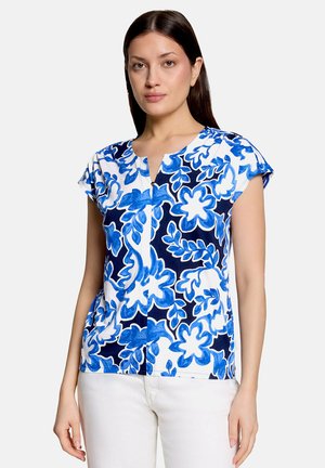 Vrouw draagt een korte mouwen witte blouse met blauwe bloemenpatronen en een witte broek, staand tegen een effen lichte achtergrond.