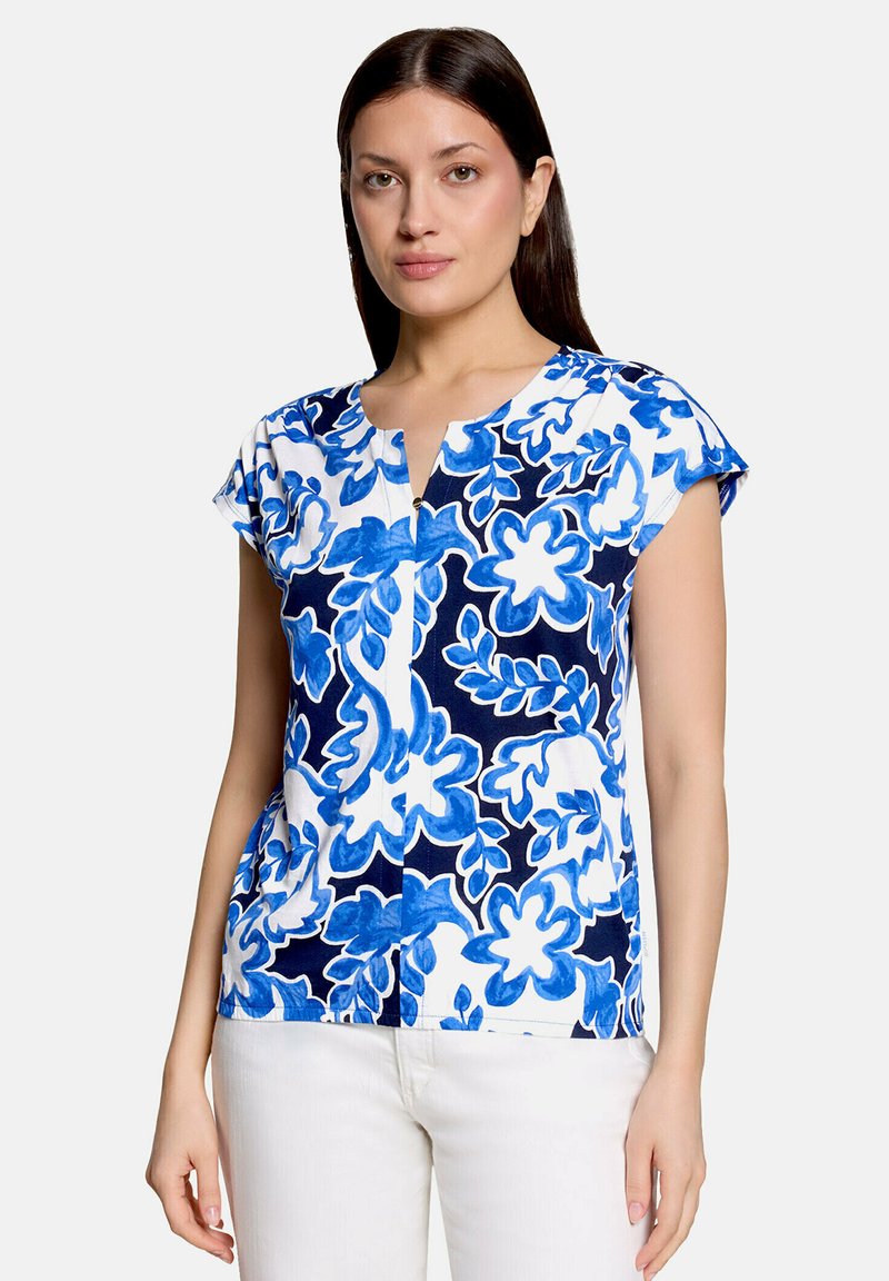 Femme portant un chemisier blanc à manches courtes avec des motifs floraux bleus et un pantalon blanc, debout devant un fond clair uni.
