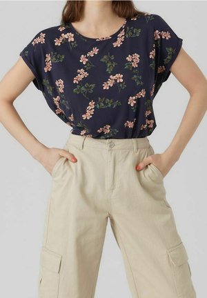 Frau trägt ein marineblaues, kurzärmeliges Oberteil mit Blumenmuster, das in hoch taillierte beige Cargohosen gesteckt ist, Hände in den Taschen.