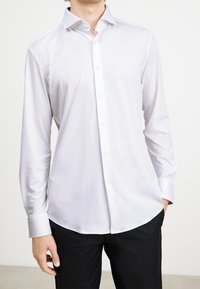 Witte blouse met lange mouwen en een puntige kraag, knoopsluiting aan de voorkant en een gladde, lichte stof. Gecombineerd met zwarte broek.