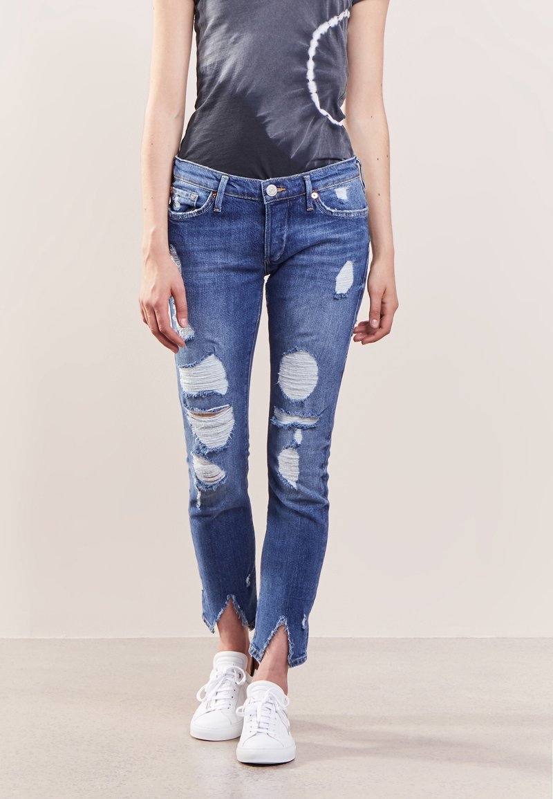 Personne portant un jean bleu très usé avec des ourlets effilochés, un t-shirt foncé et des baskets blanches, debout à l'intérieur.