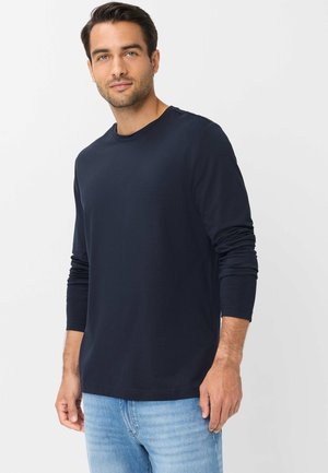 Mand med kort mørkt hår iført en marineblå langærmet skjorte og lyseblå jeans mod en ensfarvet lys baggrund.