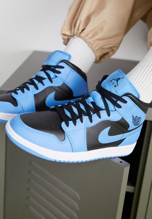 Person, die blaue und schwarze Nike Air Jordan High-Top-Sneaker mit schwarzen Schnürsenkeln, weißen Rippsocken und beigen Hosen trägt und auf einem grauen Spind sitzt.