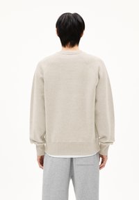 Persoon met kort zwart haar van achteren te zien, gekleed in een beige sweatshirt en lichtgrijze sweatpants tegen een witte achtergrond.