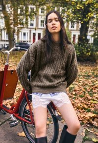 Barna kötött pulóver, fehér rövidnadrág csipkés szegéllyel, fekete térdig érő csizma, egy kerékpáron ülve egy faleveles parkban.